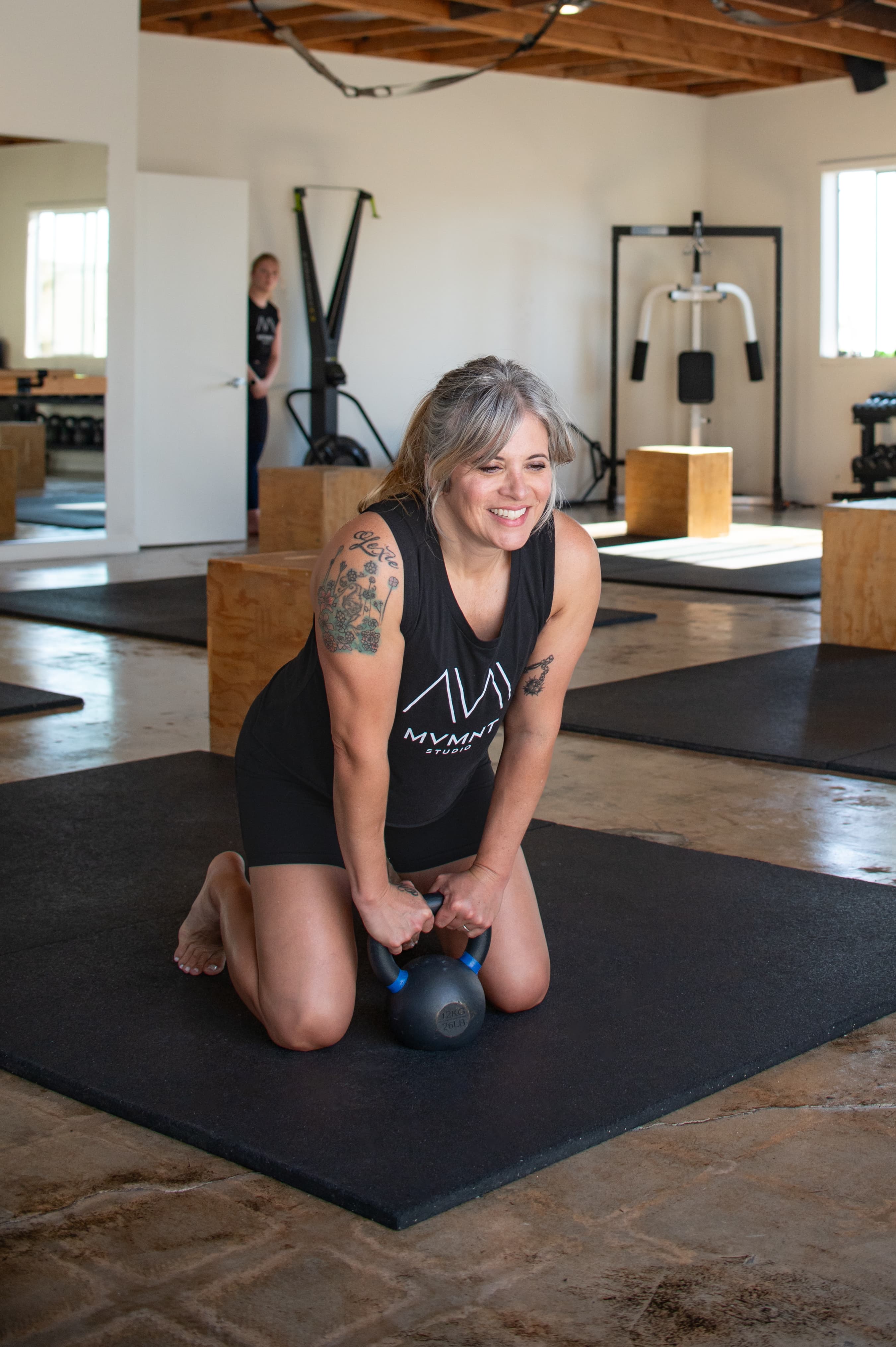 Smiling woman with tattoos kneels on a gym mat while holding a black kettlebell.